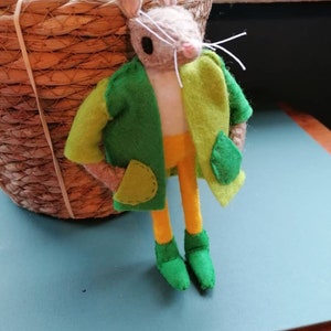 May include: A felt mouse wearing a green and yellow jacket with a yellow shirt and green shoes. The mouse is leaning against a wicker basket.