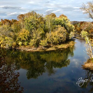 Pode incluir: Uma vista panorâmica de um rio serpenteando por uma floresta com folhagem de outono. As árvores são uma mistura de amarelo, laranja e verde. O rio está calmo e reflete o céu e as árvores. A imagem é assinada "Joyce Shelton".