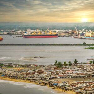 Könnte beinhalten: Luftaufnahme einer Küstenstadt bei Sonnenuntergang. Mehrere große Frachtschiffe liegen im Hafen. Ein dicht besiedeltes Gebiet mit vielen Gebäuden ist entlang der Küste zu sehen. Der Himmel ist eine Mischung aus Orange, Gelb und Blau.