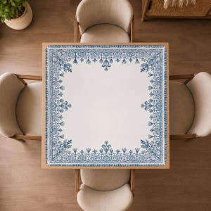 Puede incluir: Un mantel cuadrado con un centro blanco y un borde ancho e intrincado que presenta un patrón floral y de volutas en azul. El diseño incluye motivos simétricos a lo largo de los bordes y esquinas, creando un marco detallado en la tela.
