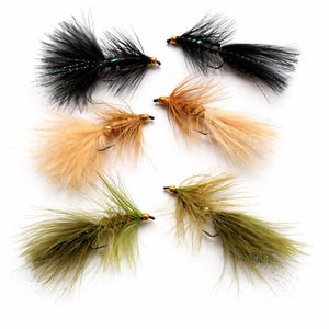 Könnte beinhalten: Sechs künstliche Angelköder in verschiedenen Farben, darunter Schwarz, Beige und Olivgrün. Jeder Köder hat einen Haken und ist mit flauschigen, federartigen Materialien versehen. Die Köder sind auf weißem Hintergrund angeordnet, um ihre komplizierten Details hervorzuheben.