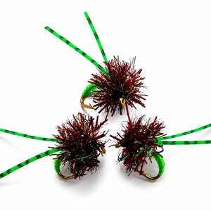 Op de afbeelding: Drie kunstmatige vliegvisvliegen. Elke vlieg heeft een donkerrood en bruin lichaam, een groene haak en groen en zwart gestreepte antennes. De vliegen zijn geplaatst tegen een witte achtergrond.