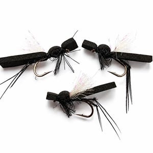 Pode incluir: Três moscas de pesca de espuma preta com anzóis metálicos, apresentando material de asa branco iridescente e caudas de fibra preta fina. Cada mosca tem uma construção de corpo segmentado com uma cabeça distinta e um dorso de espuma alongado.