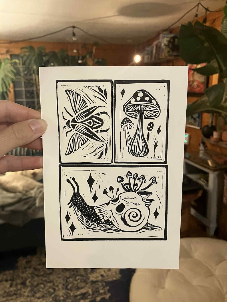 Peut inclure: Estampe en linogravure en noir et blanc avec quatre panneaux d'illustrations sur le th&egrave;me de la nature. Les panneaux repr&eacute;sentent un scarab&eacute;e, des champignons et un escargot avec un cr&acirc;ne et des champignons. L'estampe est tenue par une main.