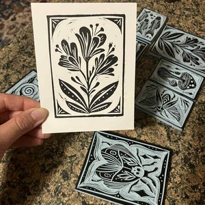 Peut inclure: Une impression linograv&eacute;e florale en noir et blanc, tenue par une main, avec plusieurs blocs de linol&eacute;um sculpt&eacute;s pr&eacute;sentant des motifs botaniques et d'insectes. L'impression montre une fleur stylis&eacute;e. Les blocs ont des fonds bleu clair.