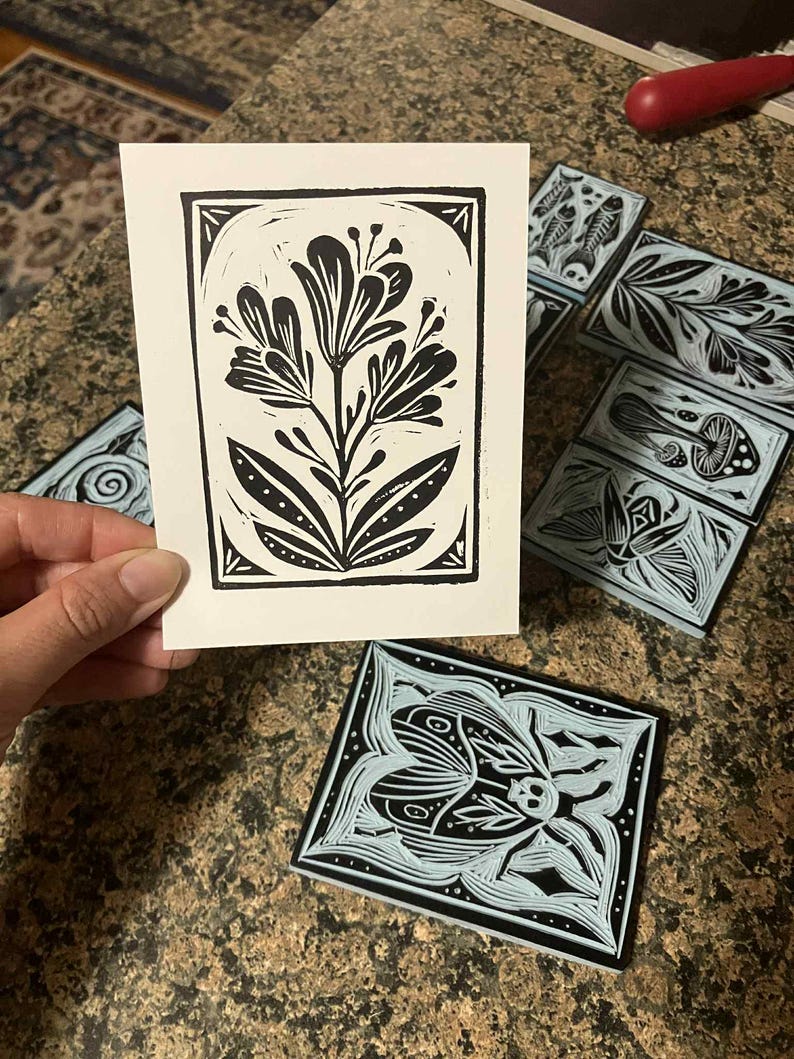 Peut inclure: Une impression linograv&eacute;e florale en noir et blanc, tenue par une main, avec plusieurs blocs de linol&eacute;um sculpt&eacute;s pr&eacute;sentant des motifs botaniques et d'insectes. L'impression montre une fleur stylis&eacute;e. Les blocs ont des fonds bleu clair.