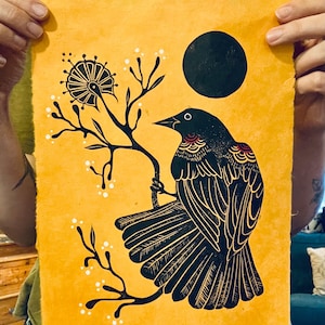 Puede incluir: Impresión de un pájaro negro sobre un fondo amarillo. El pájaro está posado en una rama con flores blancas. Un círculo negro está en la esquina superior derecha. La obra de arte está en manos de una persona.