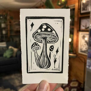Peut inclure: Estampe en linogravure en noir et blanc repr&eacute;sentant un motif de champignons. L'estampe pr&eacute;sente un grand champignon central avec un chapeau pointill&eacute;, accompagn&eacute; de petits champignons et de formes de diamants. L'&oelig;uvre est sur une carte rectangulaire blanche.