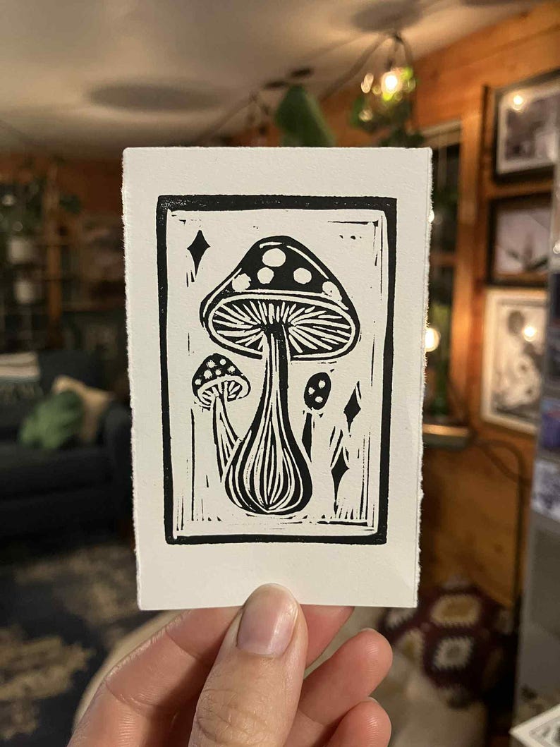 Peut inclure: Estampe en linogravure en noir et blanc repr&eacute;sentant un motif de champignons. L'estampe pr&eacute;sente un grand champignon central avec un chapeau pointill&eacute;, accompagn&eacute; de petits champignons et de formes de diamants. L'&oelig;uvre est sur une carte rectangulaire blanche.