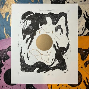 Puede incluir: Grabado en blanco y negro con dos figuras estilizadas con forma de lobo con patrones de estrellas, un círculo dorado y acentos de constelaciones. La impresión está sobre papel blanco, con fondos de papel de colores visibles.