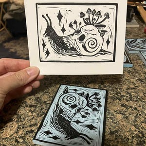 Peut inclure: Une estampe en linogravure en noir et blanc repr&eacute;sentant un escargot avec une coquille en forme de cr&acirc;ne, orn&eacute;e de champignons. L'estampe est tenue au-dessus d'un bloc de linol&eacute;um bleu et noir avec un motif similaire. Th&egrave;me gothique.