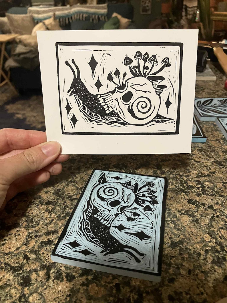 Peut inclure: Une estampe en linogravure en noir et blanc repr&eacute;sentant un escargot avec une coquille en forme de cr&acirc;ne, orn&eacute;e de champignons. L'estampe est tenue au-dessus d'un bloc de linol&eacute;um bleu et noir avec un motif similaire. Th&egrave;me gothique.