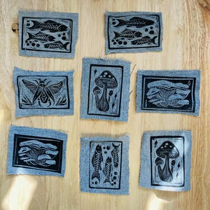 Może przedstawiać: Siedem naszywek z dżinsu z czarnymi wzorami linorytu przedstawiającymi szkielety ryb, grzyby, ćmę i rośliny. Naszywki ułożone są na jasnobrązowej drewnianej powierzchni.