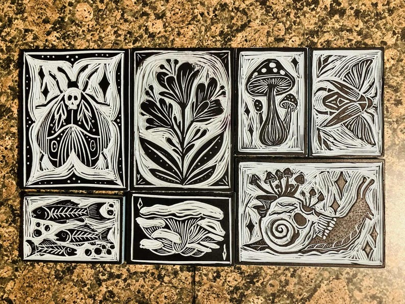 Peut inclure: Ensemble de huit gravures sur linol&eacute;um en noir et blanc, avec des motifs inspir&eacute;s de la nature. Les impressions repr&eacute;sentent un papillon de nuit, des fleurs, des champignons, un oiseau, des squelettes de poissons et un escargot. Les &oelig;uvres sont dispos&eacute;es sur une surface tachet&eacute;e brune.