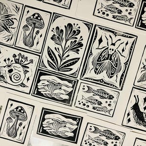Op de afbeelding: Zwart-wit linoleumsnede afdrukken met verschillende op de natuur geïnspireerde ontwerpen. De afdrukken bevatten afbeeldingen van paddenstoelen, slakken, motten, visskeletten en bloemelementen. De afdrukken zijn gerangschikt op een witte ondergrond.