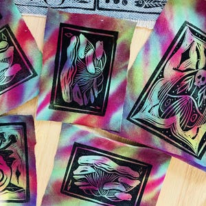 May include: A collection of fabric squares with black linocut prints. The prints feature various designs, including hands, mushrooms, and insect-like creatures. The fabric has a tie-dye effect in shades of pink, purple, green, and yellow.