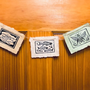 May include: A handmade banner featuring three rectangular paper prints with black linocut designs. The prints depict a mushroom, fish skeletons, and a moth. The banner is strung on a thin twine and hangs against a wooden door.