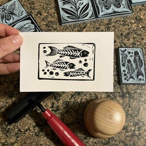 Peut inclure: Une estampe en linogravure en noir et blanc de deux squelettes de poissons et de bulles sur papier blanc. L'estampe est tenue pr&egrave;s de plusieurs blocs de linol&eacute;um sculpt&eacute;s avec des motifs floraux et d'insectes. Une roulette &agrave; manche rouge et un brunissoir en bois sont &eacute;galement visibles.