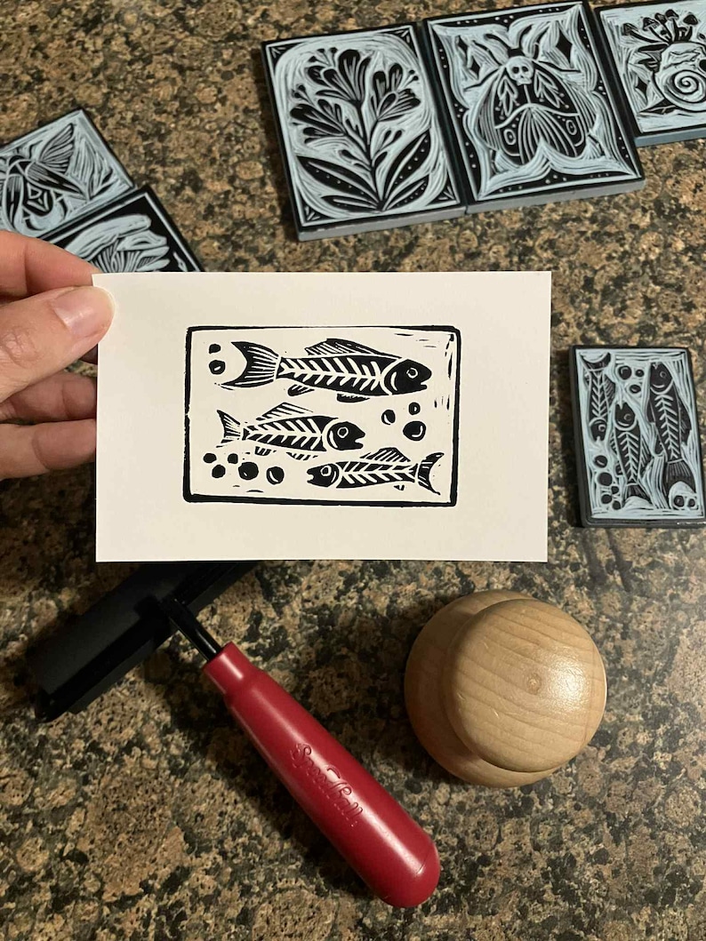 Peut inclure: Une estampe en linogravure en noir et blanc de deux squelettes de poissons et de bulles sur papier blanc. L'estampe est tenue pr&egrave;s de plusieurs blocs de linol&eacute;um sculpt&eacute;s avec des motifs floraux et d'insectes. Une roulette &agrave; manche rouge et un brunissoir en bois sont &eacute;galement visibles.