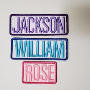May include: Three embroidered name patches. The top patch reads "JACKSON" in purple thread on a white background, framed by a purple border. The middle patch says "WILLIAM" in blue thread with a blue border. The bottom patch says "ROSE" in pink thread with a pink border.