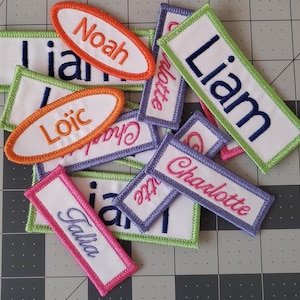 May include: A collection of colourful embroidered fabric name tags with various names including Liam, Noah, Loic, Charlotte, and Faisa.