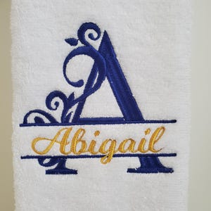 May include: White bath towel with a blue embroidered letter A with a gold floral design and the name Abigail embroidered below.