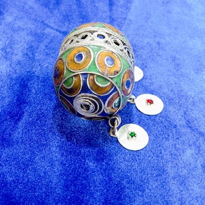 Pode incluir: Um ornamento em forma de ovo de prata e esmalte com um design decorativo de círculos verdes, azuis e laranja. O ornamento tem três pingentes de prata com pedras vermelhas e verdes.
