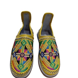 Puede incluir: Un par de zapatos sin cordones amarillos con coloridos patrones geométricos bordados. El diseño incluye triángulos, diamantes y zigzags en varios colores, incluyendo verde, azul, naranja y morado. Los zapatos tienen una suela blanca.
