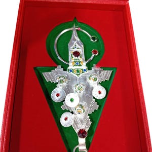 Peut inclure: Pendentif argenté finement détaillé, présenté sur un triangle de velours vert dans un coffret rouge. Le pendentif comporte des pierres précieuses colorées et un porte-clés. L'intérieur du coffret est en velours vert.
