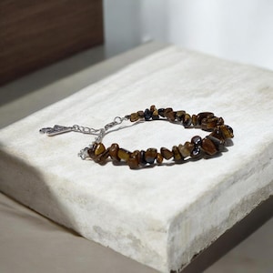 Peut inclure: Un bracelet composé de petites pierres d'œil de tigre de forme irrégulière, dans des tons bruns et dorés. Le bracelet est doté d'une chaîne et d'un fermoir argentés. Les pierres sont assemblées pour former un cercle complet.