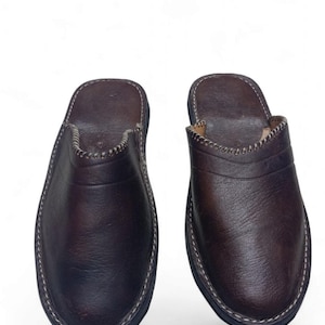 Puede incluir: Un par de zapatillas de cuero marrón oscuro. Las zapatillas tienen una puntera redondeada, un borde blanco cosido y una suela oscura. El cuero parece liso con sutiles variaciones de color. El diseño es sencillo y clásico.