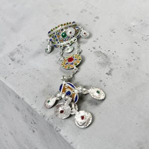 Peut inclure: Une broche en métal argenté ornée d'un travail d'émail complexe dans des tons bleu, vert, orange et blanc. La pièce se compose d'une section supérieure décorative reliée par des chaînes à un pendentif inférieur, avec plusieurs breloques.