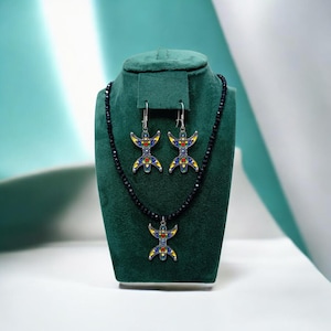 Peut inclure: Un ensemble de bijoux composé d'un collier et de boucles d'oreilles assorties. Le collier présente une chaîne de perles noires avec un pendentif argenté. Les boucles d'oreilles sont des pendentifs argentés avec des détails en émail bleu, jaune et rouge.