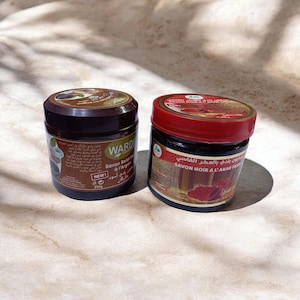 May include: Two jars of Wardi brand Savon Beldi soap. One jar has a brown lid and label with text, the other has a red lid and label with text. The jars are on a light brown surface.