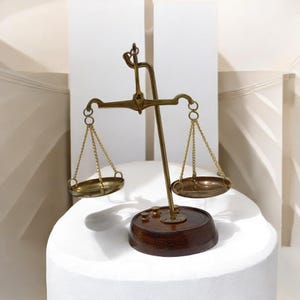 May include: An antique brass balance scale with two suspended pans, each linked by chains. The scale is set on a dark wooden base. The background is a neutral white, emphasizing the vintage design of the weighing scale.