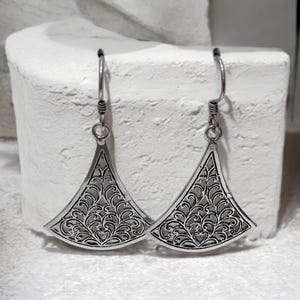 May include: A pair of silver-toned earrings with a triangular shape and intricate filigree design. The earrings have a hook closure and a detailed, openwork pattern. The design features a repeating floral and heart motif, creating a delicate and ornate appearance.