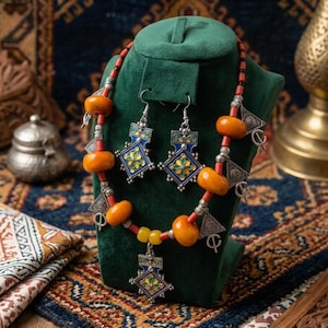 Peut inclure: Un ensemble collier et boucles d'oreilles composé de perles orange et jaunes, d'accents en métal argenté et de pendentifs en émail bleu avec des motifs floraux jaunes et verts. L'ensemble est présenté sur un support en velours vert.