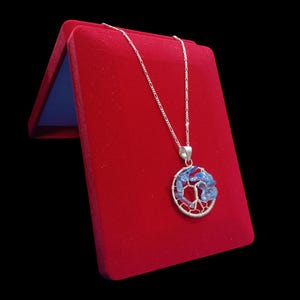 Peut inclure: Un collier en argent avec un pendentif en forme d'arbre rempli de pierres bleues. Le pendentif est suspendu à une fine chaîne en argent. Le collier est présenté sur un écrin en velours rouge sur fond noir.