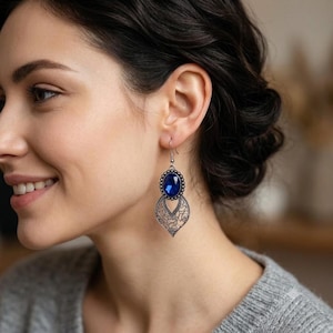 Peut inclure: Une boucle d'oreille pendante composée d'une pierre ovale bleu profond sertie dans un cadre argenté festonné. Sous la pierre se trouve un élément en métal filigrané en forme de goutte avec des motifs ajourés, suspendu à un crochet argenté.