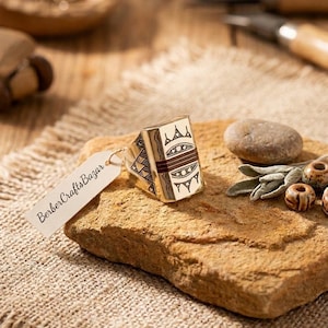 Peut inclure: Une bague dorée avec des motifs géométriques et une face rectangulaire. La bague est présentée sur une surface en pierre avec une étiquette indiquant "BerberCraftsBazar". La bague présente un design détaillé avec des accents marron et blancs.