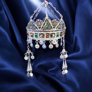 Puede incluir: Una corona plateada con intrincados detalles de filigrana y esmalte colorido. La corona presenta detalles en rojo, verde y azul, con colgantes y una delicada cadena. El fondo es una tela azul oscuro.