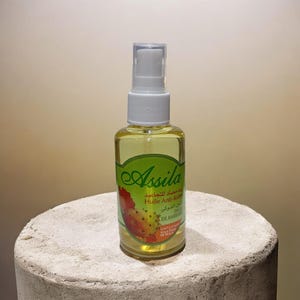 Peut inclure: Une bouteille en verre transparente avec un bouchon pulvérisateur blanc, remplie de liquide jaune. La bouteille porte une étiquette verte avec le mot "Assila" en cursive et du texte en plusieurs langues. La bouteille est posée sur une surface claire et texturée.