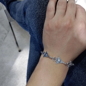 Peut inclure: Bracelet en argent orné de breloques triangulaires ornées de motifs complexes. Le bracelet est relié par de petits connecteurs circulaires. Le poignet est mis en valeur sur un fond de jean bleu.