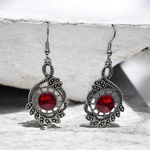 Peut inclure: Une paire de boucles d'oreilles pendantes argentées. Chaque boucle d'oreille présente une pierre rouge ronde au centre, entourée d'un motif ajouré orné. Le design comprend un motif floral et une boucle décorative en haut, avec un crochet pour oreilles percées.