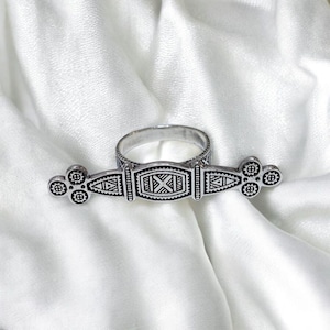 Peut inclure: Une bague argentée au design orné. La bague présente une pièce maîtresse rectangulaire avec un motif en forme de X et des détails géométriques. La bande de la bague est reliée à une barre avec des accents circulaires et triangulaires.
