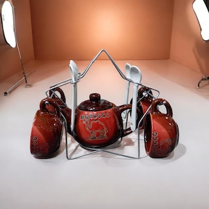 Peut inclure: Un service à thé en céramique comprenant une théière orange foncé et marron avec un motif de chameau et le mot MOROCCO, accompagné de tasses assorties et de cuillères blanches, le tout maintenu dans un support en fil métallique argenté.