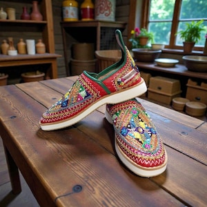 May include: A pair of red slip-on shoes with intricate embroidery in various colors, including green, yellow, and blue. The shoes have a white sole and a green accent on the heel. The design features geometric patterns and floral motifs.