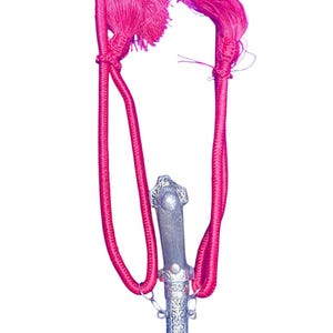 Puede incluir: Una daga decorativa con una hoja curva y un mango plateado adornado. La daga está suspendida por un cordón rosa brillante con borlas a juego. El mango presenta diseños intrincados y la hoja tiene una ligera curva.