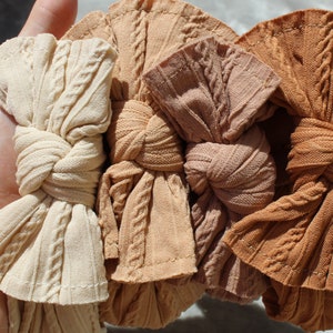 May include: Four knotted headbands in shades of beige and brown. Each headband has a cable knit pattern and a knotted bow. The headbands are made from a soft, textured fabric, suitable for babies or children.
