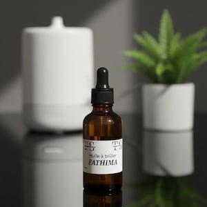 Könnte beinhalten: Eine braune Glasflasche mit schwarzer Pipette, beschriftet mit "Huile à brûler Fathima". Im Hintergrund ein weißer Diffusor und eine kleine Topfpflanze. Die Flasche spiegelt sich auf einer glänzenden schwarzen Oberfläche.